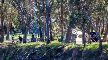 Family campsite beside the Murray River with a large beige tent, campfire setup, and people gathered among tall gum trees under dappled sunlight.