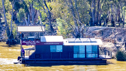 Compact modern houseboat named Houseboat for Two moored on the Murray River, with solar panels on the roof and two people aboard surrounded by gum trees.
