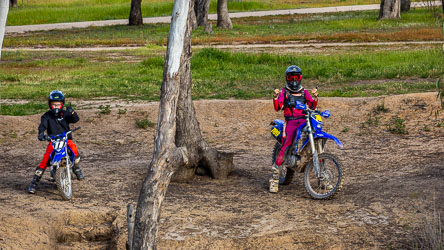Two youngsters wearing protective motocross gear and helmets on blue dirt bikes, posing on a sandy riverbank with gum trees and grassy background.