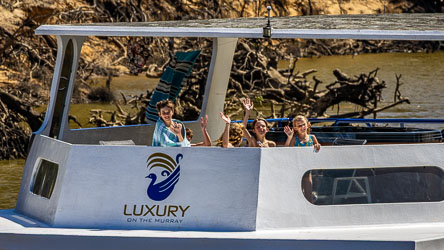 Family waving from the deck of a white houseboat named Luxury on the Murray, with towels hanging behind and gum‑lined riverbank in the background.