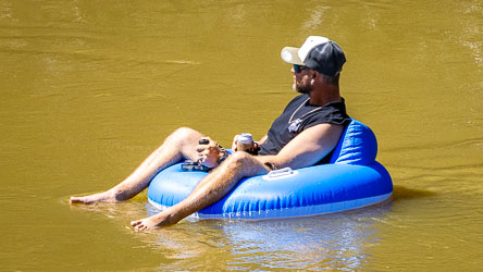 Man relaxing on a bright blue inflatable tube floating on the Murray River, wearing sunglasses and a cap, holding a drink and enjoying the sunshine.