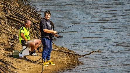 Two men fishing from a sandy riverbank near Echuca, one seated on a cooler and the other standing with a rod, surrounded by tree roots and calm water.