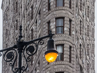 Close‑up of the Flatiron Building in New York City showing ornate stone façade and windows with a vintage streetlamp glowing in the foreground.