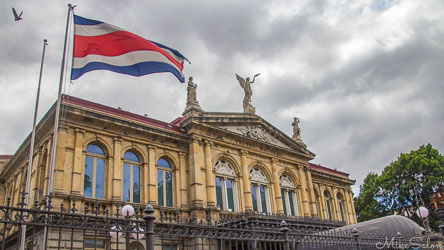 Front view of the National Theatre in San José, Costa Rica, showing its ornate neoclassical façade with statues and arched windows beneath a cloudy sky, the Costa Rican flag flying prominently at left.