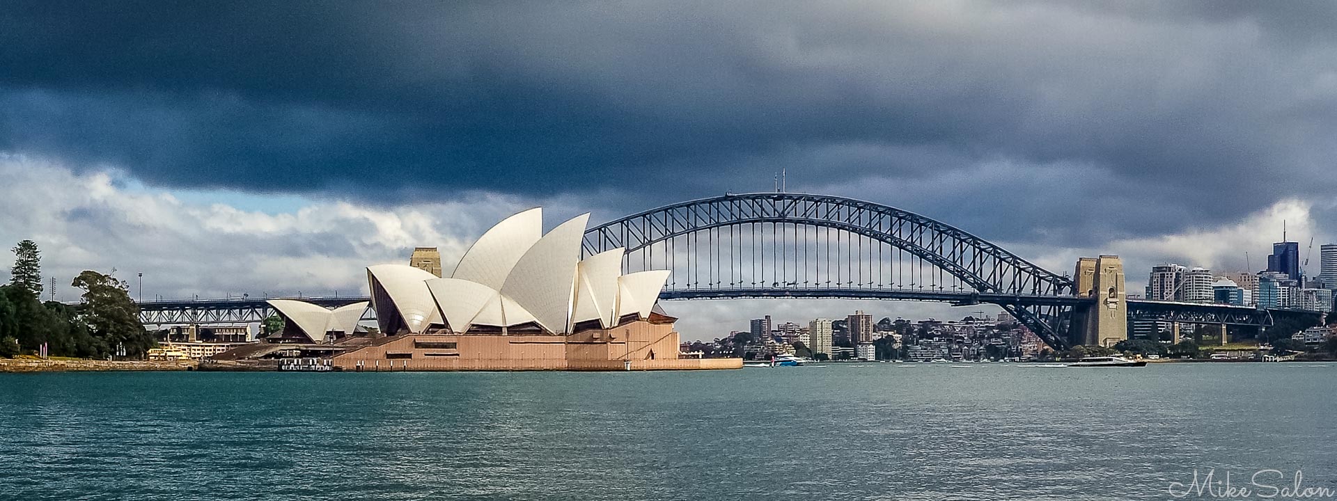 A dramatic perspective of Sydney Harbour, showcasing two of Australia's most recognized architectural landmarks framed against a brooding sky. In the foreground, the gleaming white sails of Jørn Utzon