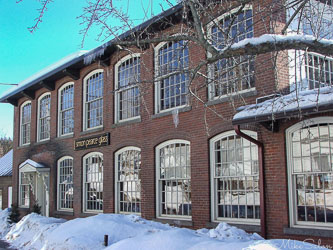 Brick building housing Simon Pearce glassworks in Quechee, Vermont, with large arched windows and snow on the ground and roof.