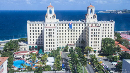 Aerial view of Hotel Nacional de Cuba in Havana with twin towers, palm‑lined gardens, swimming pool, and blue sea in the background.