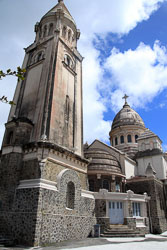 View from low on the hillside of Sacré‑Cœur de Balata in Martinique, showing its pale domed church façade against a blue sky with scattered clouds.