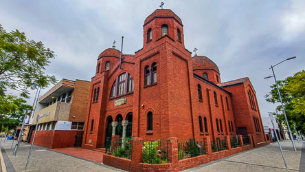 Red brick Greek Orthodox church in Byzantine Revival style on a city street corner, featuring twin domed towers, arched windows, and a central tiled dome against a cloudy sky.